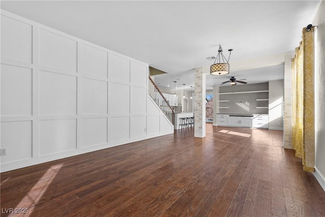 unfurnished living room with stairway, dark wood-style flooring, a ceiling fan, and a decorative wall