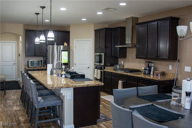 kitchen with visible vents, light stone counters, a kitchen breakfast bar, stainless steel appliances, and wall chimney range hood