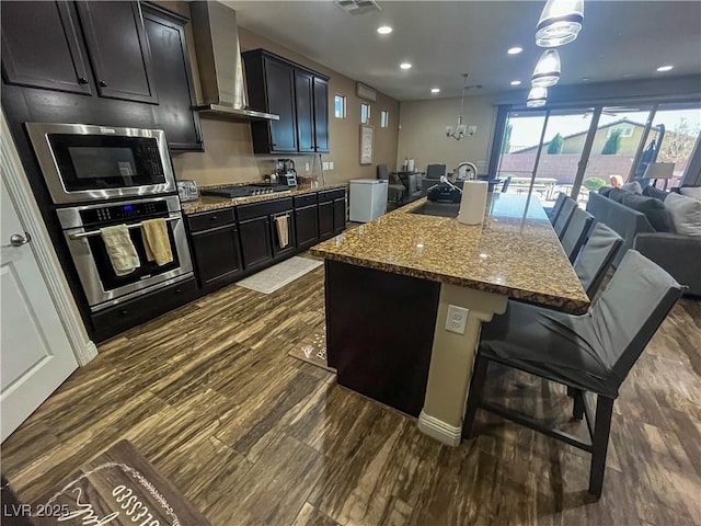 kitchen with a sink, dark stone countertops, open floor plan, stainless steel appliances, and wall chimney range hood