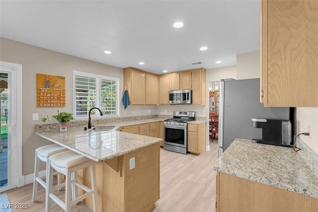 kitchen with light brown cabinets, a sink, light stone counters, appliances with stainless steel finishes, and a peninsula