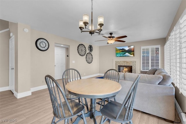 dining space with a glass covered fireplace, ceiling fan with notable chandelier, baseboards, and light wood-style floors