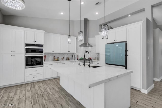 kitchen with stainless steel double oven, an inviting chandelier, freestanding refrigerator, and wall chimney range hood