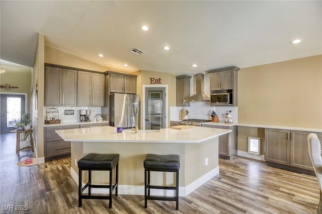 kitchen featuring a center island with sink, vaulted ceiling, wood finished floors, stainless steel appliances, and wall chimney exhaust hood