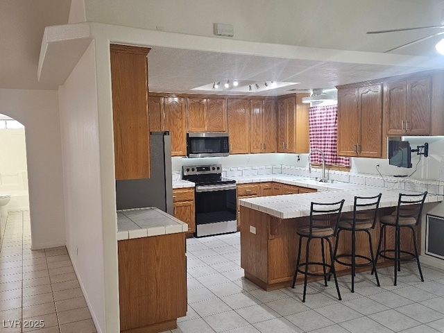 kitchen with a sink, a peninsula, brown cabinets, stainless steel appliances, and light tile patterned flooring