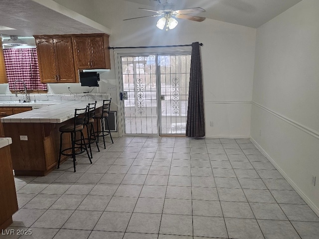 kitchen with light tile patterned floors, a sink, a ceiling fan, a breakfast bar, and tile countertops