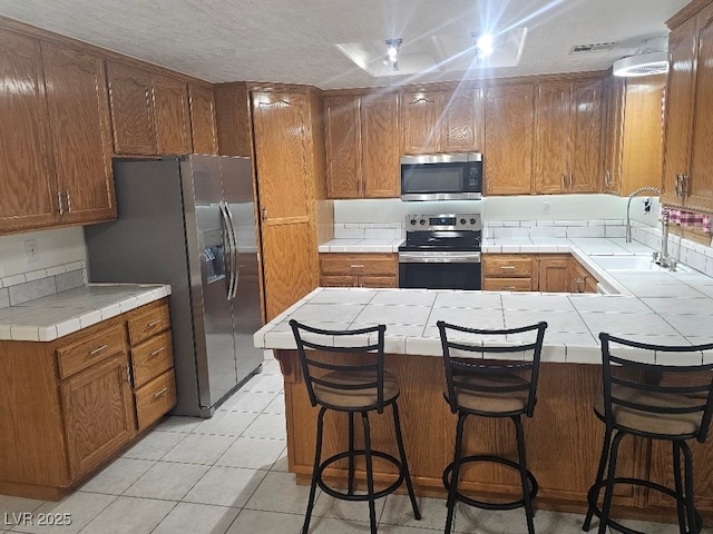 kitchen with a peninsula, light tile patterned flooring, a sink, stainless steel appliances, and tile counters