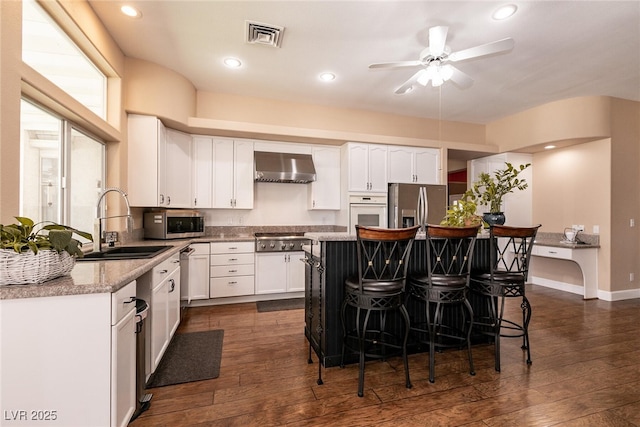 kitchen featuring a sink, wall chimney range hood, appliances with stainless steel finishes, a ceiling fan, and a breakfast bar area