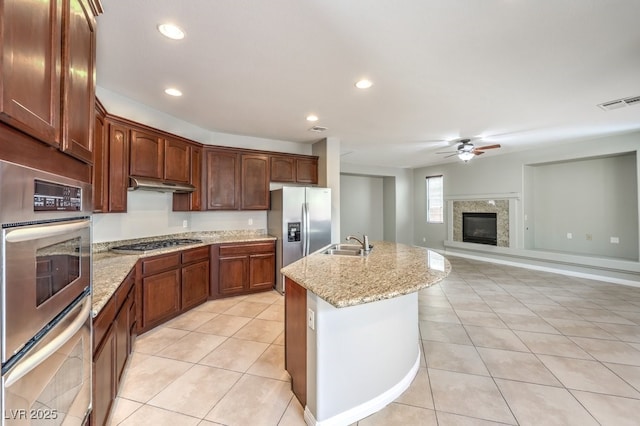 kitchen with stainless steel appliances, a glass covered fireplace, a sink, ceiling fan, and an island with sink