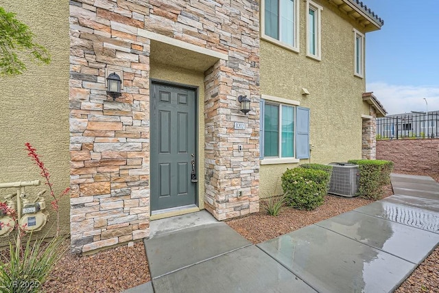 doorway to property featuring stone siding, central AC unit, stucco siding, and fence