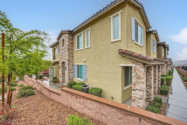view of side of home featuring central air condition unit, fence, stone siding, stucco siding, and a tiled roof