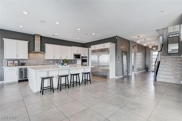 kitchen with light countertops, a kitchen island with sink, wall chimney exhaust hood, beverage cooler, and built in appliances