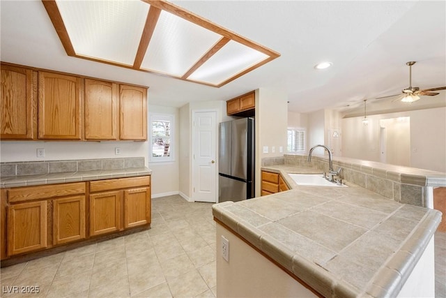 kitchen featuring a sink, freestanding refrigerator, tile counters, a peninsula, and ceiling fan