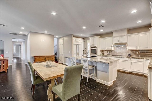 kitchen with white cabinets, under cabinet range hood, dark wood-style flooring, and an island with sink