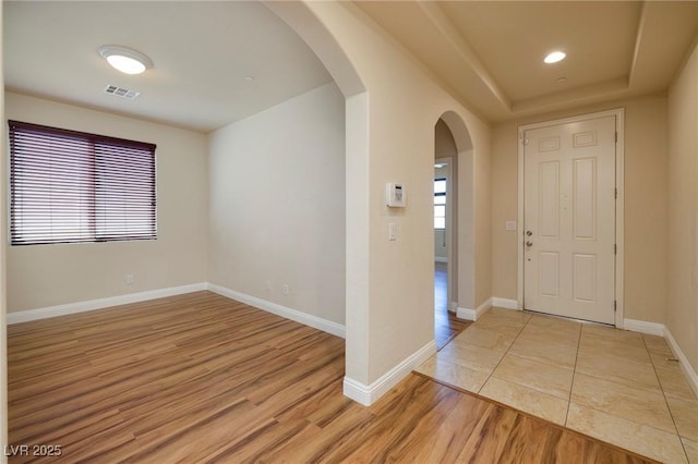 foyer featuring visible vents, baseboards, a tray ceiling, arched walkways, and light wood-style flooring