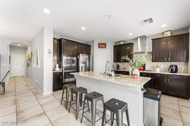 kitchen with a breakfast bar area, wall chimney exhaust hood, appliances with stainless steel finishes, visible vents, and arched walkways