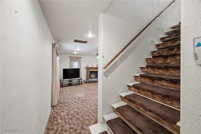 stairs featuring a tiled fireplace, carpet flooring, a ceiling fan, visible vents, and a textured wall