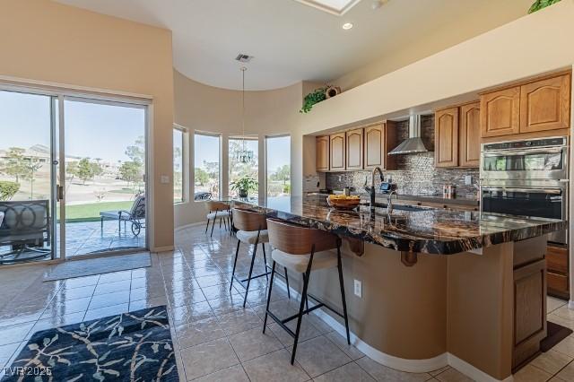 10524 Longwood Dr, Las Vegas, NV 89134 - kitchen featuring a breakfast bar area, decorative backsplash, dark stone counters, double oven, and wall chimney exhaust hood