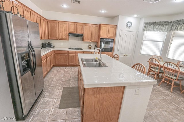 kitchen with oven, tile countertops, an island with sink, a sink, and stainless steel fridge with ice dispenser