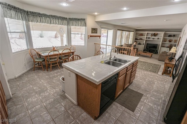 kitchen with black dishwasher, a sink, tile counters, brown cabinetry, and a kitchen island with sink