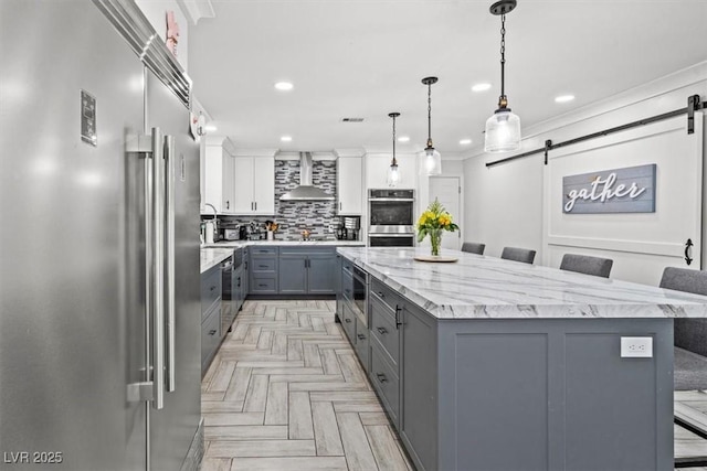 kitchen with wall chimney range hood, a spacious island, a barn door, gray cabinetry, and stainless steel appliances