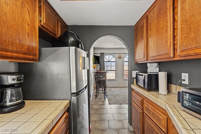 kitchen with brown cabinetry, black microwave, arched walkways, freestanding refrigerator, and tile counters