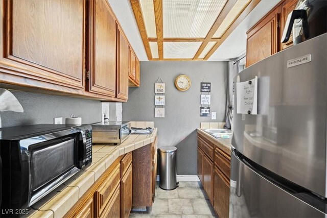 kitchen with freestanding refrigerator, baseboards, stone finish floor, tile countertops, and brown cabinets