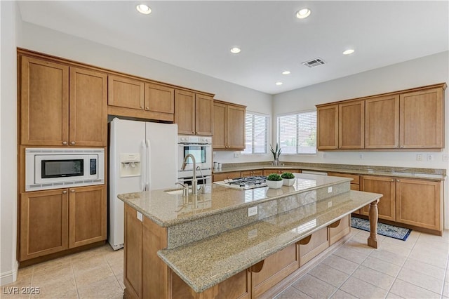 kitchen featuring a sink, brown cabinetry, white appliances, and a center island with sink