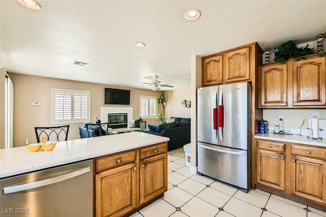 kitchen with light countertops, brown cabinets, stainless steel appliances, a ceiling fan, and a glass covered fireplace