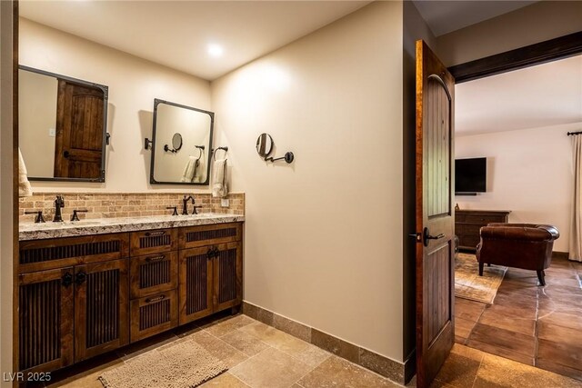 bathroom featuring baseboards, backsplash, double vanity, and stone tile flooring