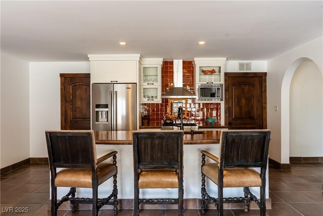 kitchen featuring stainless steel appliances, wall chimney exhaust hood, arched walkways, a kitchen bar, and glass insert cabinets