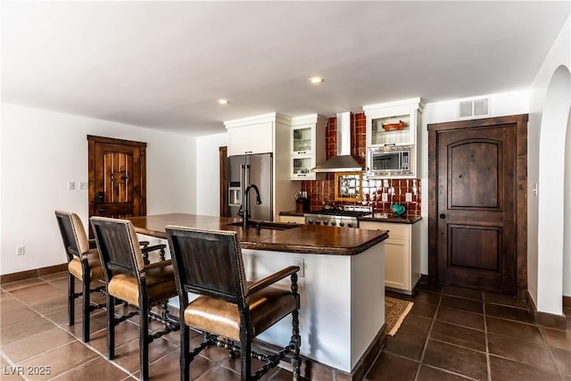 kitchen with a sink, wall chimney range hood, dark countertops, appliances with stainless steel finishes, and a breakfast bar area