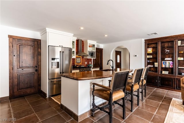 kitchen featuring stainless steel appliances, wall chimney exhaust hood, arched walkways, dark countertops, and a sink