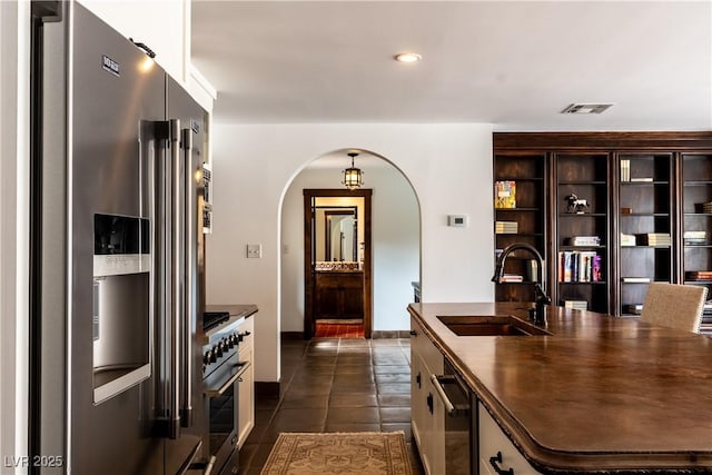 kitchen with premium appliances, a sink, arched walkways, dark tile patterned floors, and a kitchen island with sink
