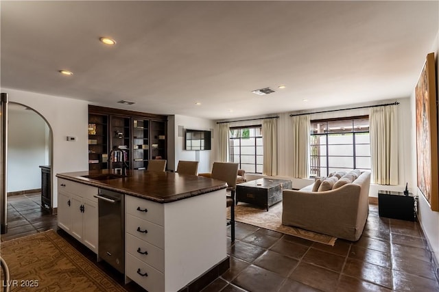 kitchen with a sink, dark countertops, white cabinetry, arched walkways, and a center island with sink
