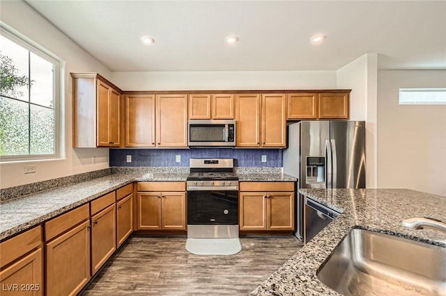 kitchen with a sink, dark wood-style flooring, brown cabinetry, stainless steel appliances, and stone counters