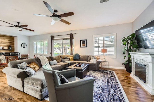 living area with a glass covered fireplace, baseboards, a ceiling fan, visible vents, and wood finished floors