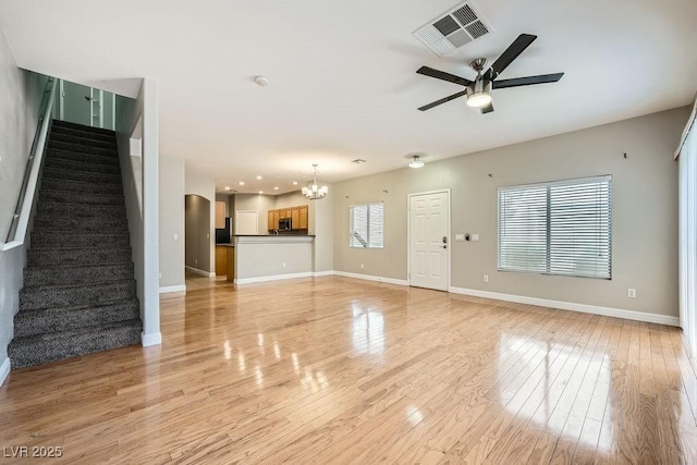 unfurnished living room featuring baseboards, stairway, light wood-style flooring, visible vents, and ceiling fan with notable chandelier