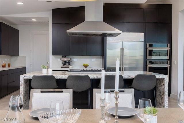 kitchen featuring dark cabinets, modern cabinets, a kitchen breakfast bar, wall chimney range hood, and appliances with stainless steel finishes