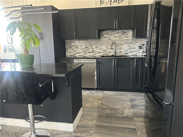kitchen featuring dark stone counters, a sink, dishwasher, dark cabinets, and freestanding refrigerator