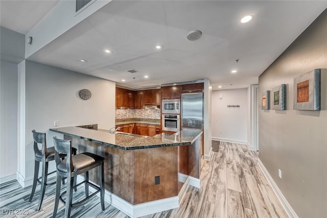 kitchen with built in appliances, dark stone counters, a peninsula, baseboards, and light wood-type flooring