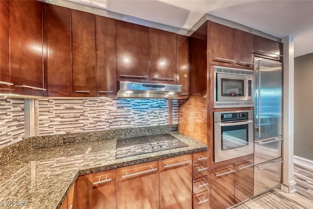 kitchen with decorative backsplash, baseboards, dark stone countertops, built in appliances, and under cabinet range hood