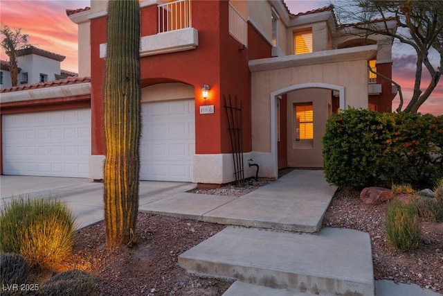 exterior entry at dusk featuring concrete driveway, a balcony, stucco siding, an attached garage, and a tiled roof