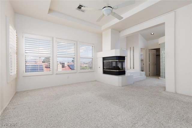 unfurnished living room with ceiling fan, carpet, a multi sided fireplace, visible vents, and a raised ceiling