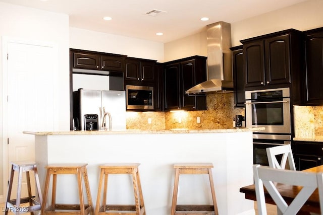 kitchen featuring wall chimney exhaust hood, stainless steel appliances, tasteful backsplash, visible vents, and a kitchen bar