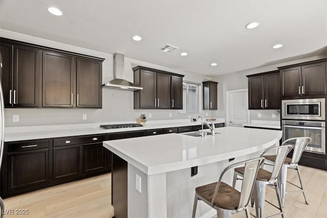 kitchen featuring appliances with stainless steel finishes, wall chimney exhaust hood, a sink, a kitchen bar, and an island with sink