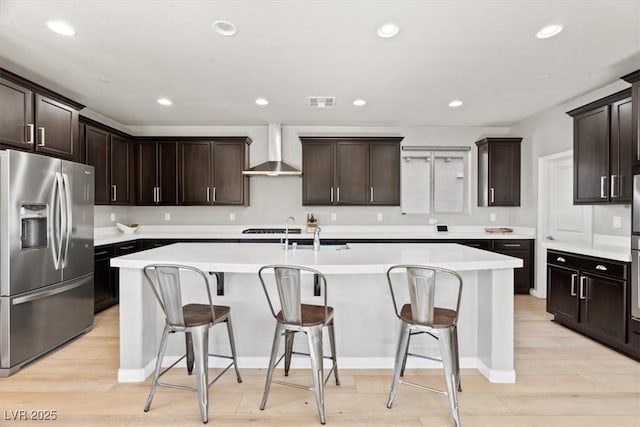 kitchen with stainless steel fridge with ice dispenser, wall chimney exhaust hood, a center island with sink, a breakfast bar, and light wood-style floors