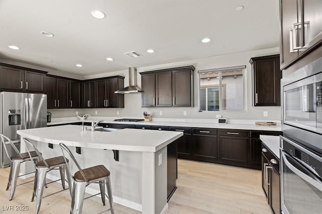 kitchen with stainless steel appliances, wall chimney exhaust hood, a breakfast bar, a kitchen island with sink, and light wood finished floors
