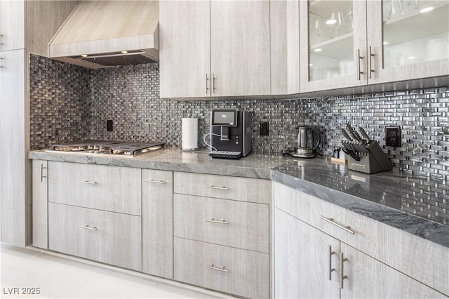 kitchen featuring premium range hood, stainless steel gas stovetop, tasteful backsplash, light stone countertops, and glass insert cabinets