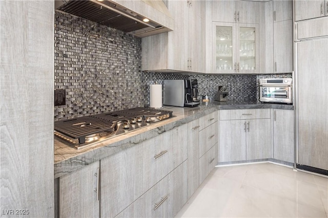 kitchen with wall chimney range hood, decorative backsplash, stainless steel gas stovetop, and glass insert cabinets