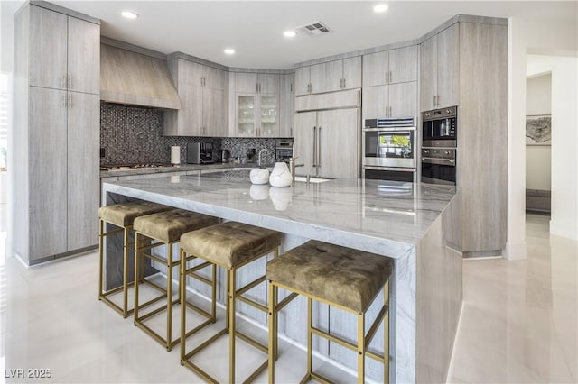 8000 Clock Tower Ct, Las Vegas, NV 89117 - kitchen with light stone countertops, decorative backsplash, a kitchen bar, custom exhaust hood, and a large island with sink